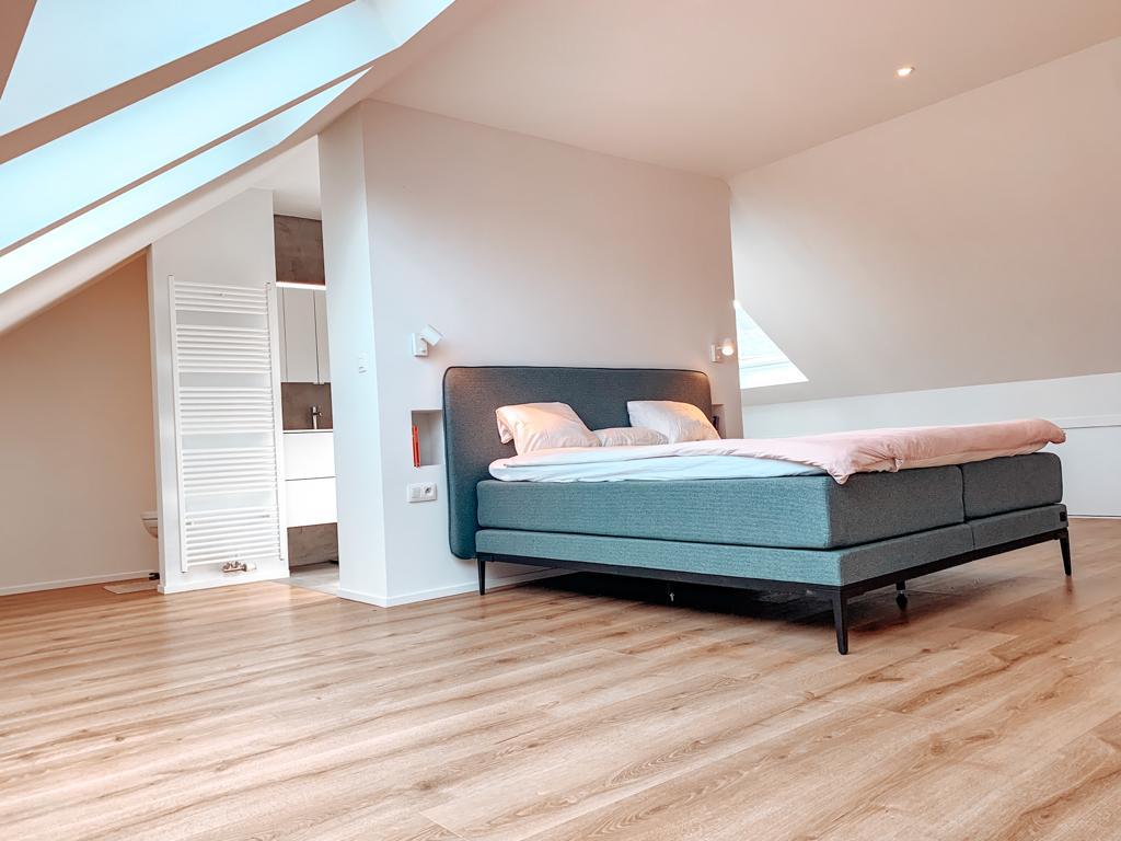 Moderne master bedroom onder een schuin dak met grote raampartijen, afgewerkt met een strakke houten vloer en een open naadloze overgang naar de badkamer.